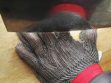 A kitchen knife is cutting back of chainmail glove on the chopping board.